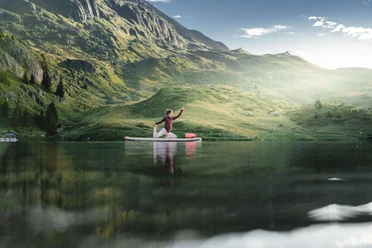 SUP Yoga auf dem Bettmersee Frau übt SUP Yoga Bettmersee im Sommer auf einem ruhigen Bergsee umgeben von grünen Hängen und alpiner Landschaft