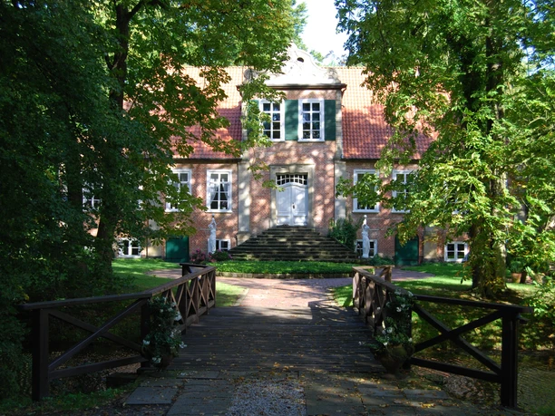 Fikensolt2.JPG Das Bild zeigt ein historisches Anwesen mit einer eleganten Fassade, die von üppigem Grün umrahmt wird. Eine hölzerne Brücke führt über einen kleinen Graben zum Eingang des Gebäudes, dessen Treppe zur Haustür aus dunklem Stein besteht.