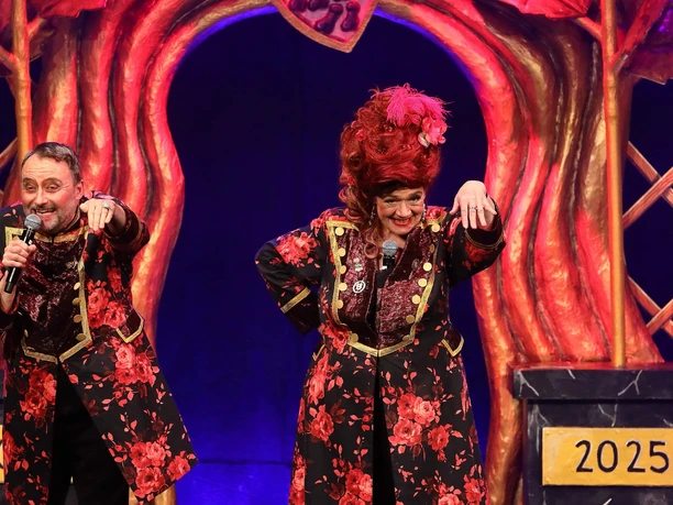 Röschen Sitzung in Cologne Two people stand on a stage with red, floral costumes in front of an artistically designed background.