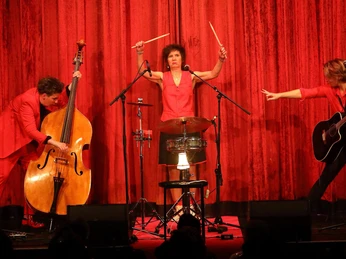 Röschen Sitzung in Cologne Drei Personen in roten Outfits auf der Bühne mit Bass, Schlagzeug und Gitarre vor rotem Vorhang.Three people in red outfits on stage with bass, drums and guitar in front of a red curtain.
