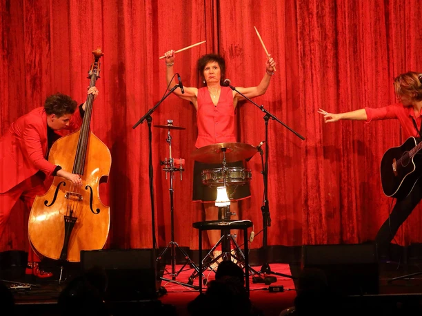 Röschen Sitzung in Cologne Three people in red outfits on stage with bass, drums and guitar in front of a red curtain.