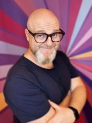 Glenn Langhorst - Photo by VEEDELIA.jpg Mann mit Glatze und Brille vor buntem Strahlenhintergrund, lächelt mit leicht geneigtem Kopf.