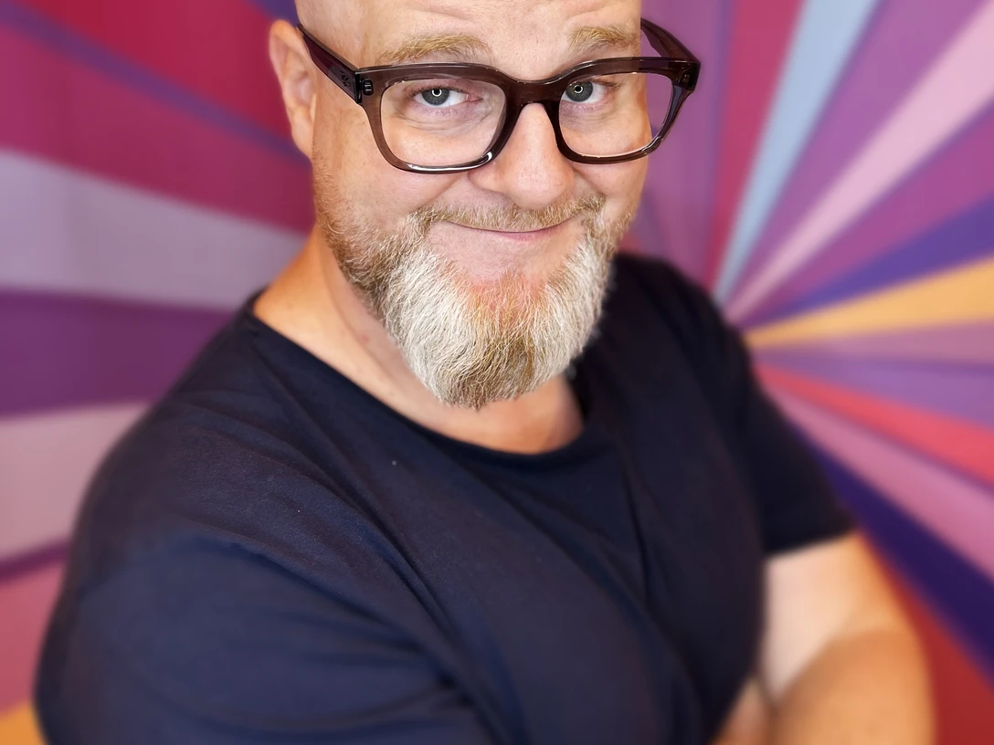 Glenn Langhorst - Photo by VEEDELIA.jpg Mann mit Glatze und Brille vor buntem Strahlenhintergrund, lächelt mit leicht geneigtem Kopf.