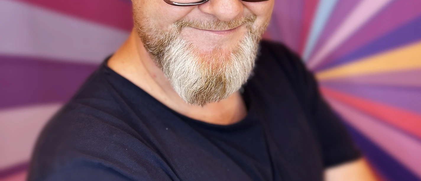 Glenn Langhorst - Photo by VEEDELIA.jpg Mann mit Glatze und Brille vor buntem Strahlenhintergrund, lächelt mit leicht geneigtem Kopf.