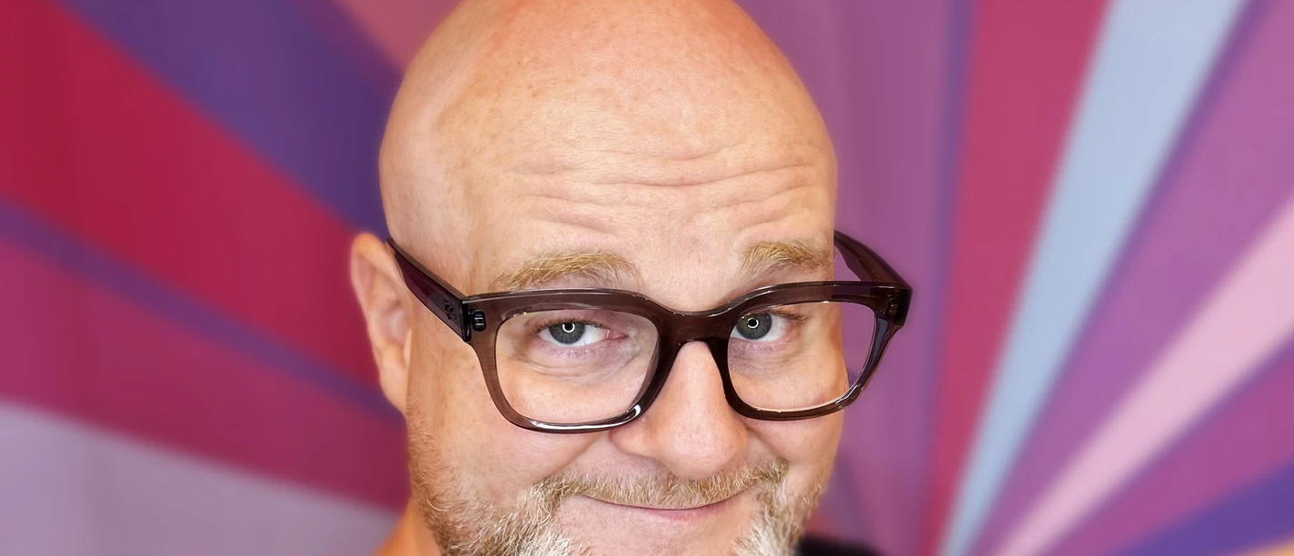 Glenn Langhorst - Photo by VEEDELIA.jpg Mann mit Glatze und Brille vor buntem Strahlenhintergrund, lächelt mit leicht geneigtem Kopf.