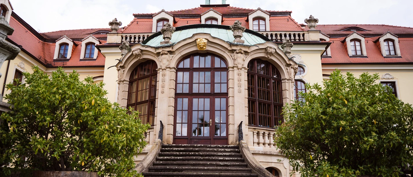 Schloss Proschwitz.jpg Historisches Schloss mit rotem Dach, Turmspitze und kunstvollen Steindetails, flankiert von Pflanzen.