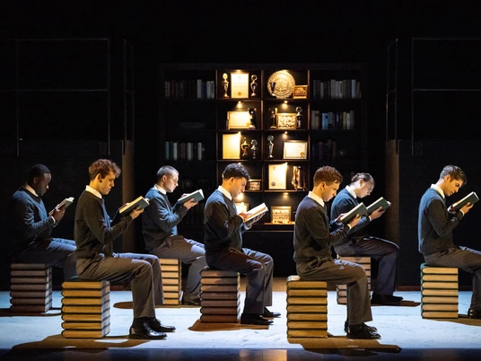 Der Club der toten Dichter.jpg Das Bild zeigt sieben Schüler, die jeweils akkurat auf einem Holzhocker in einer Bibliothek sitzen und Bücher lesen.