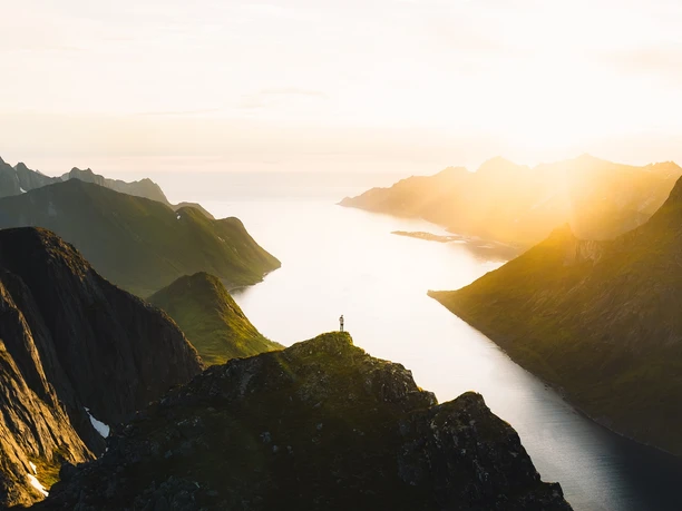 Norwegen Felsige Berglandschaft im Vordergrund, dahinter ein einsamer Wanderer, mit Blick auf das sonnenbeschienene Fjord.
