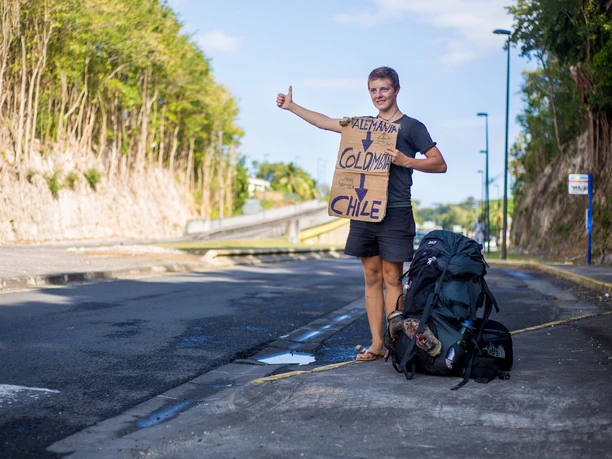 Hitchhiking to Tierra del Fuego Young man standing at the side of the road, thumbs up, holding sign: Trip through Colombia, Chile planned.
