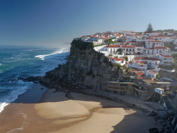 Portugal & Madeira Ein Küstenort an einer Klippe mit weißen Häusern, der über einem Strand mit sanften Wellen thront.