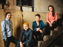 Vier Personen sitzen auf einer Treppe in einem Industriegebäude, lässig posierend.