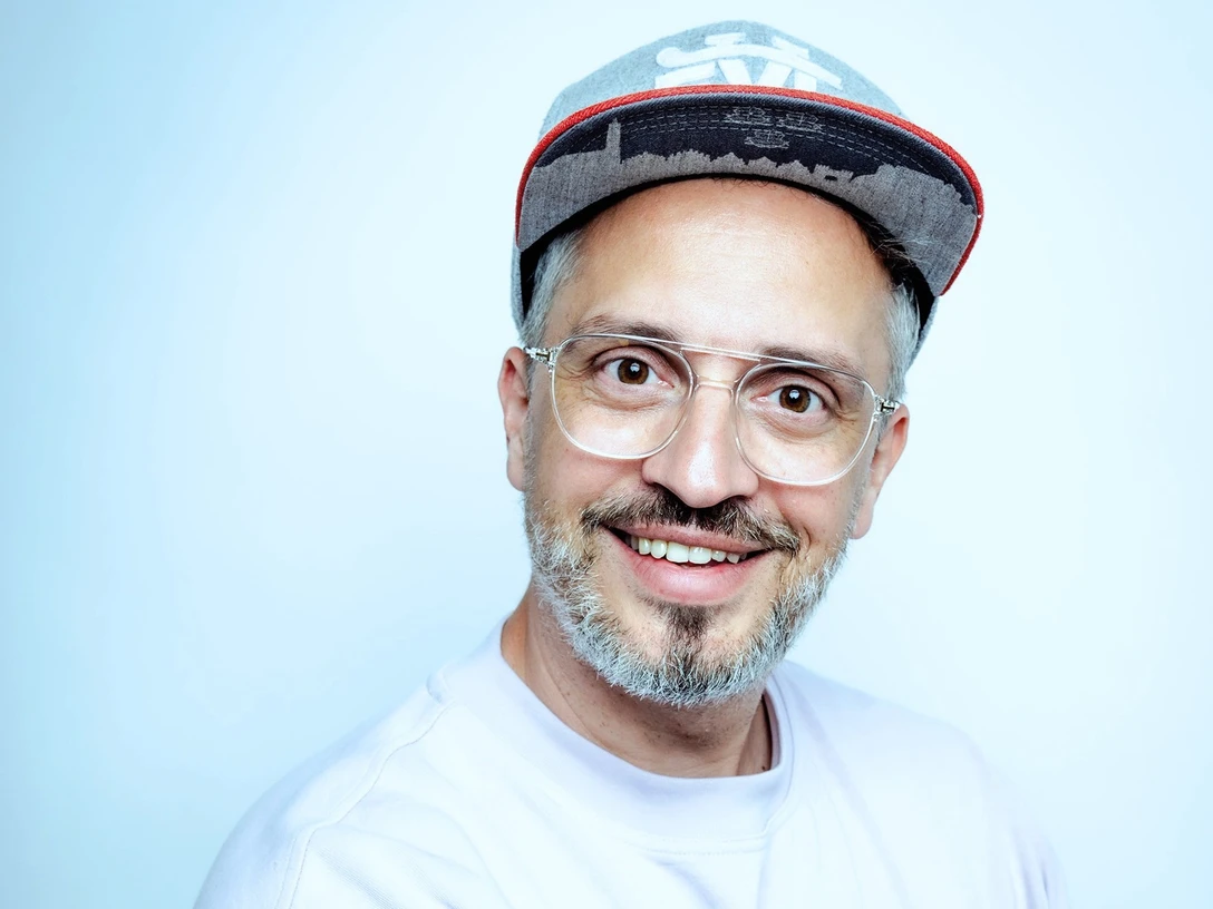 derphysiopath-claudius-pflugcmyk_quer.jpg Mann mit Brille und Basecap lächelt freundlich vor hellblauem Hintergrund.