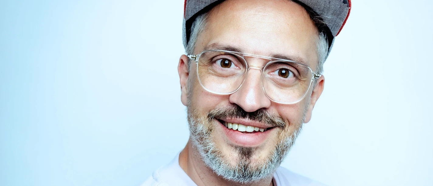 derphysiopath-claudius-pflugcmyk_quer.jpg Mann mit Brille und Basecap lächelt freundlich vor hellblauem Hintergrund.