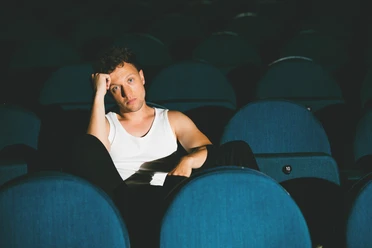 joris-c-luis-jantsch-cmyk-quer (1).jpg Person in weißem Tanktop sitzt nachdenklich zwischen leeren blauen Kinosesseln.