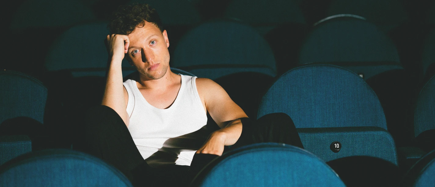 joris-c-luis-jantsch-cmyk-quer (1).jpg Person in weißem Tanktop sitzt nachdenklich zwischen leeren blauen Kinosesseln.