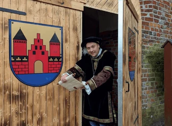 Mann in historischer Kleidung öffnet Holztor mit Stadtwappen und hält eine Schriftrolle in der Hand.
