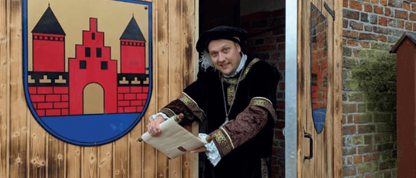 Mann in historischer Kleidung öffnet Holztor mit Stadtwappen und hält eine Schriftrolle in der Hand.