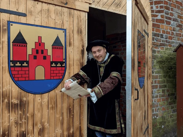 Markteröffnung.png Mann in historischer Kleidung öffnet Holztor mit Stadtwappen und hält eine Schriftrolle in der Hand.