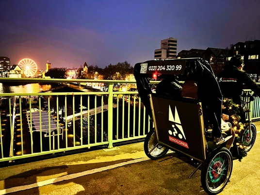 RikoloniaWinterTeamlicht.png Ein winterlich beleuchtetes Rikscha-Team vor dem Kölner Rheinpanorama bei Abenddämmerung.A wintery illuminated rickshaw team in front of the Cologne Rhine panorama at dusk.