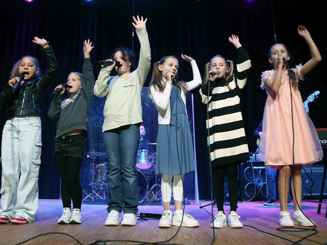 Kinder auf einer Bühne singen in Mikrofone, dahinter Musikinstrumente sichtbar. Bunte Bühnenbeleuchtung.