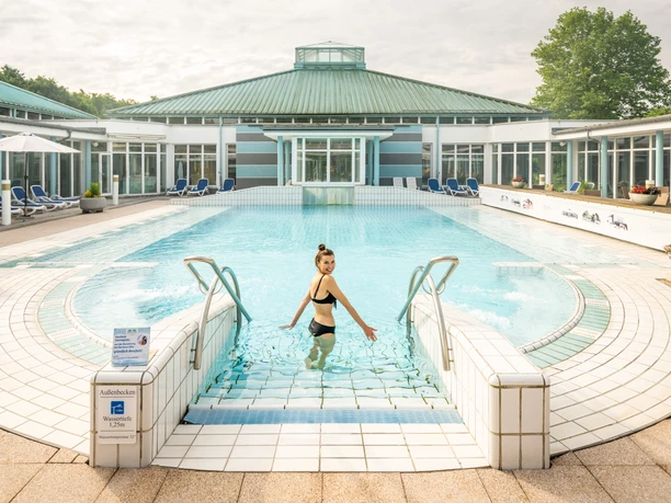 Frau im Bikini steigt lächelnd über Treppen ins Außenbecken der Driburg Therme in Bad Driburg.