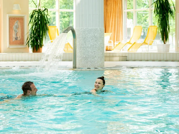 Zwei Personen entspannen im warmen Innenbecken der Driburg Therme unter einem Wasserstrahl.