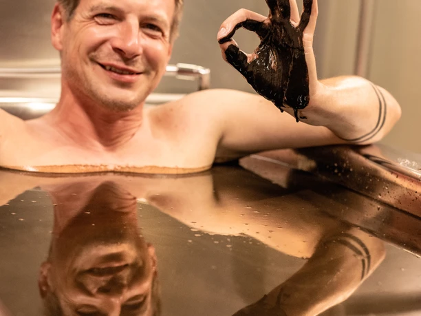 Bad-Driburg-Graefliches-Gesundheitszentrum-Moorbad-Teutoburger-Wald-Bad-Driburger-Touristik-GmbH-D-Ketz-179.jpg Mann in einem Moorbad hebt lächelnd eine mit Moor bedeckte Hand und zeigt ein Kreiszeichen.