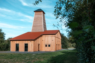 Saline Salzderhelden ©Florian Spieker Holzverkleidetes Saline mit rotem Satteldach und hohem Turm, umgeben von Bäumen und Wiese.