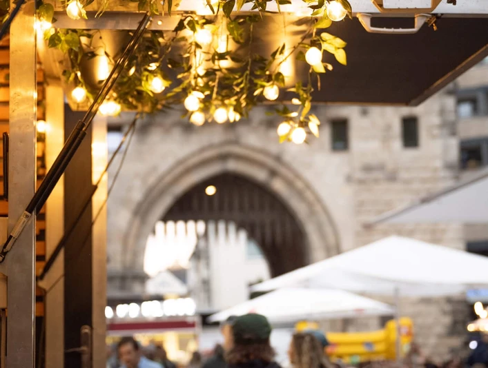 Feierabendmarkt auf dem Chlodwigplatz Ein belebter Marktplatz bei Dämmerung, mit Lichterketten und einem historischen Torbogen im Hintergrund.