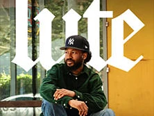 Ein Mann sitzt in lässiger Kleidung, entspannt vor einem Gebäude mit großen Fenstern.