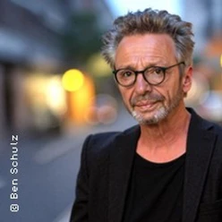 Ein Mann mit Brille und grauem Haar steht selbstbewusst in einer städtischen Umgebung.