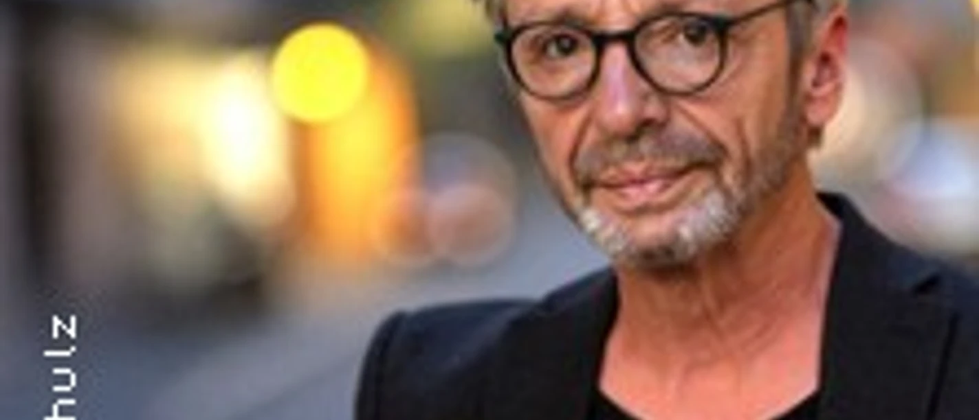 Ein Mann mit Brille und grauem Haar steht selbstbewusst in einer städtischen Umgebung.