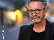 Ein Mann mit Brille und grauem Haar steht selbstbewusst in einer städtischen Umgebung.