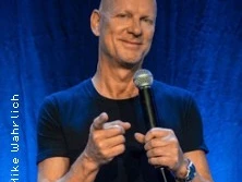 Portrait des lächelnden Mannes mit Mikrofon auf einer Bühne, vor blauem Hintergrund.