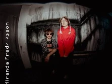 Zwei Personen posieren lächelnd, eine in einem roten Pullover, die andere in Sonnenbrille.