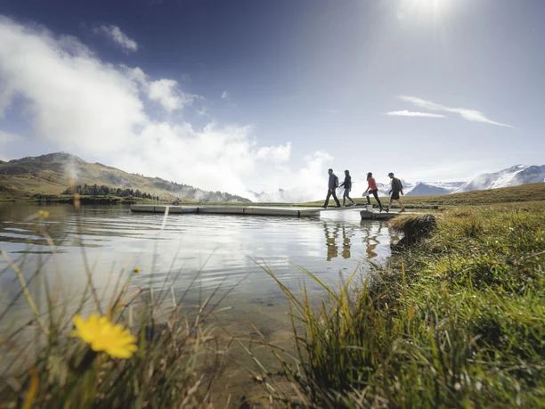 Foxtrail Wanderung am Seeufer Foxtrail Desoria Aletsch Arena mit vier Personen, die über einen Steg am Bettmersee wandern, vor den Walliser Alpen