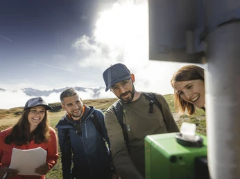 Team spirit at Foxtrail Desoria Foxtrail Desoria Aletsch Arena mit vier Teilnehmern, die gemeinsam eine Aufgabe an einer grünen Box lösenFoxtrail Desoria Aletsch Arena with four participants working together to solve a task at a green boxFoxtrail Desoria Aletsch Arena avec quatre participants qui résolvent ensemble une tâche sur une boîte verte