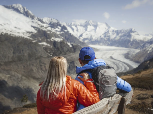 Aussicht von der Hohfluh Frühlingsfahrten Hohfluh mit einem Paar in bunten Jacken auf einer Bank mit Blick auf den Grossen Aletschgletscher