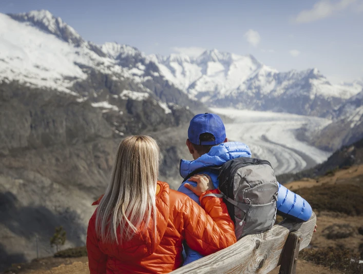 View from the Hohfluh Frühlingsfahrten Hohfluh mit einem Paar in bunten Jacken auf einer Bank mit Blick auf den Grossen AletschgletscherSpring rides Hohfluh with a couple in colorful jackets on a bench with a view of the Great Aletsch GlacierCourses printanières à Hohfluh avec un couple en vestes colorées sur un banc avec vue sur le grand glacier d'Aletsch