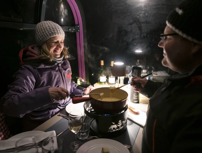 Genussvolles Fondueerlebnis im Winter Fonduegondel Bettmerhorn mit Paar beim Käsefondue in stimmungsvoller Gondel am Abend in der verschneiten Aletsch ArenaBettmerhorn fondue gondola with couple enjoying a cheese fondue in an atmospheric gondola in the snowy Aletsch Arena in the eveningTélécabine pour fondue Bettmerhorn avec couple lors d'une fondue au fromage dans une télécabine à l'ambiance chaleureuse le soir dans l'Aletsch Arena enneigée