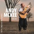 Der Musiker Andy McKee steht mit zwei Gitarren auf einem Feld bei Sonnenuntergang.