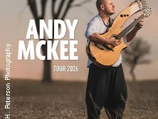 Der Musiker Andy McKee steht mit zwei Gitarren auf einem Feld bei Sonnenuntergang.