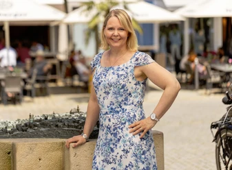 Die starken Frauen von Günzburg Stadtführerin im blauen Kleid, lächelnd, neben einem Stadtmodell, vor einem belebten Restaurant.
