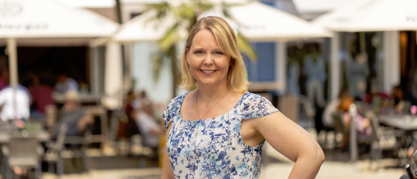Die starken Frauen von Günzburg Stadtführerin im blauen Kleid, lächelnd, neben einem Stadtmodell, vor einem belebten Restaurant.