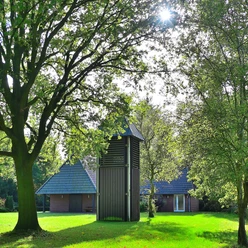 Kleine Kapelle mit dunklem Holzturm, umgeben von grüner Landschaft unter blauem Himmel.