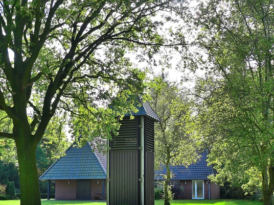 Kleine Kapelle mit dunklem Holzturm, umgeben von grüner Landschaft unter blauem Himmel.