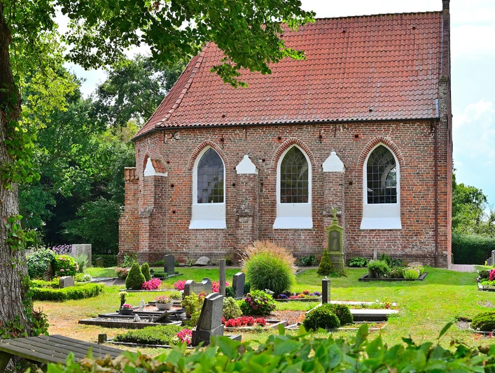 Die Kapelle in Vreschen-Bokel aus Backstein mit roten Dachziegeln, umgeben von gepflegten Gräbern und Pflanzen.