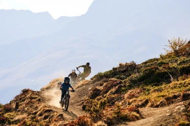 Action-packed trail ride in the Upper Valais Upper Valais bike pass with bikers on a dusty single trail in Bellwald in late summer light and against a mountain backdrop