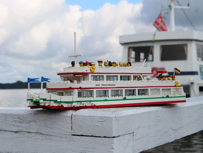 lego-weisse-flotte Modellschiff aus LEGO mit Aufschrift „Bad Zwischenahn“ vor echtem Fahrgastschiff am Seeufer