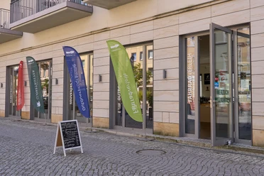 Aktivzentrum Touristinformation Bad Schandau Bunte Werbefahnen vor einem Eingang mit Tafel auf Gehweg.